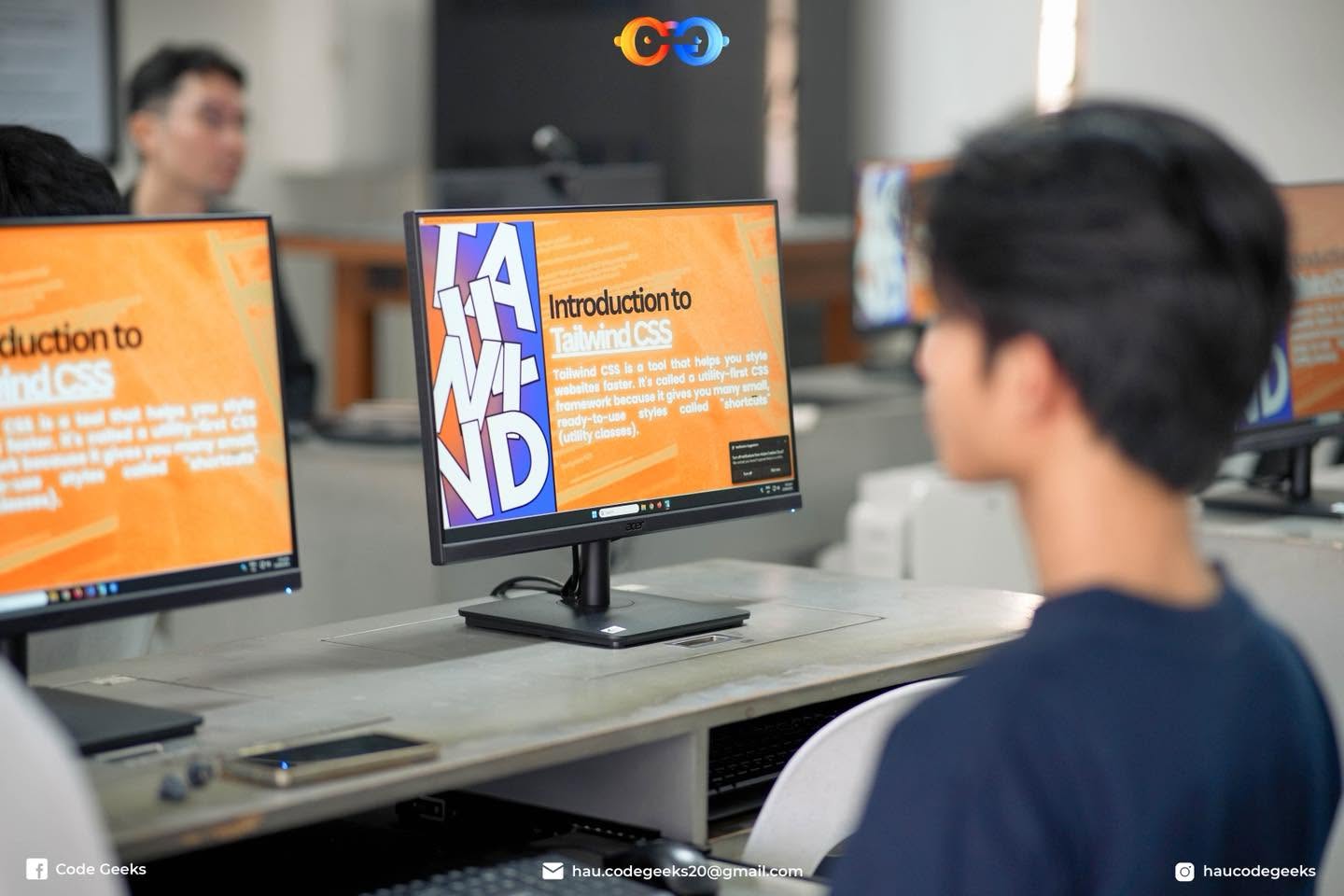 A student watching the Introduction to Tailwind CSS slide on his own monitor, the same slide mirrored down the row of machines behind him
